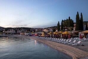 A beach with sun loungers and umbrellas, a calm sea, and buildings in the background. - Apartments Rozankovic (Okrug)
