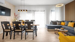 A modern living room with a sofa, dining table, and chairs. There is a TV mounted on the wall and a window with curtains. - Villa Tria Quercus (Vodice)