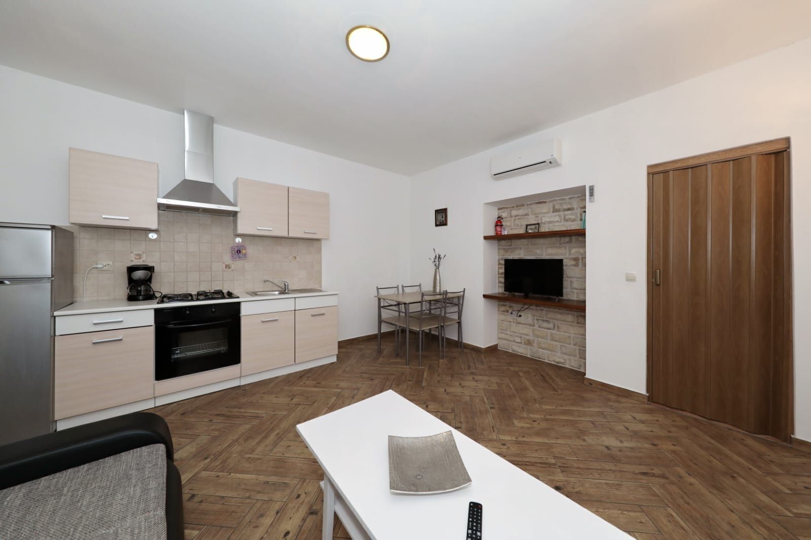 A compact kitchen with a stove, oven, and modern cabinetry. There is a dining area with a table and chairs, and a TV mounted on a stone wall.