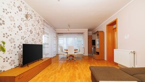 A modern living room with a flat-screen TV, wooden flooring, a dining area with a white table and chairs, and a kitchen area with wooden cabinets. - Apartment Ereiz (Crikvenica)
