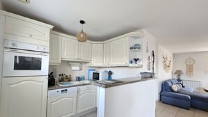 Eine Küche mit weißen Schränken, einem eingebauten Backofen, einer Mikrowelle und einem Geschirrspüler. Im angrenzenden Wohnbereich befindet sich ein blaues Sofa.