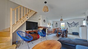 Ein geräumiges Wohnzimmer mit einer Holztreppe, einem Fernseher, blauen Sesseln und einem Essbereich mit Tisch und Stühlen.