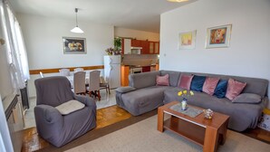 A living room with a grey sofa, a matching armchair, and a wooden coffee table. There is a dining area with a white table and chairs, and a kitchen in the background. - Apartments Jasmine (Biograd na Moru)