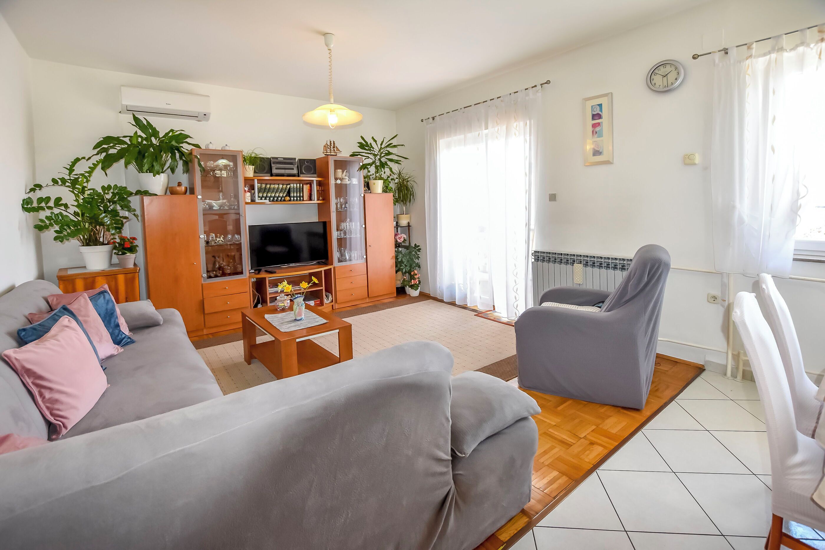 A living room with a sofa, armchair, TV, wooden cabinet, and potted plants.