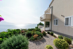Ein Gebäude mit Balkon und Blick auf ein Gewässer sowie ein angelegter Garten mit verschiedenen Pflanzen.
