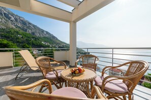 A balcony with wicker furniture and a table with fruit, overlooking a mountainous landscape and a body of water. - Apartments Olive sea View (Omis)