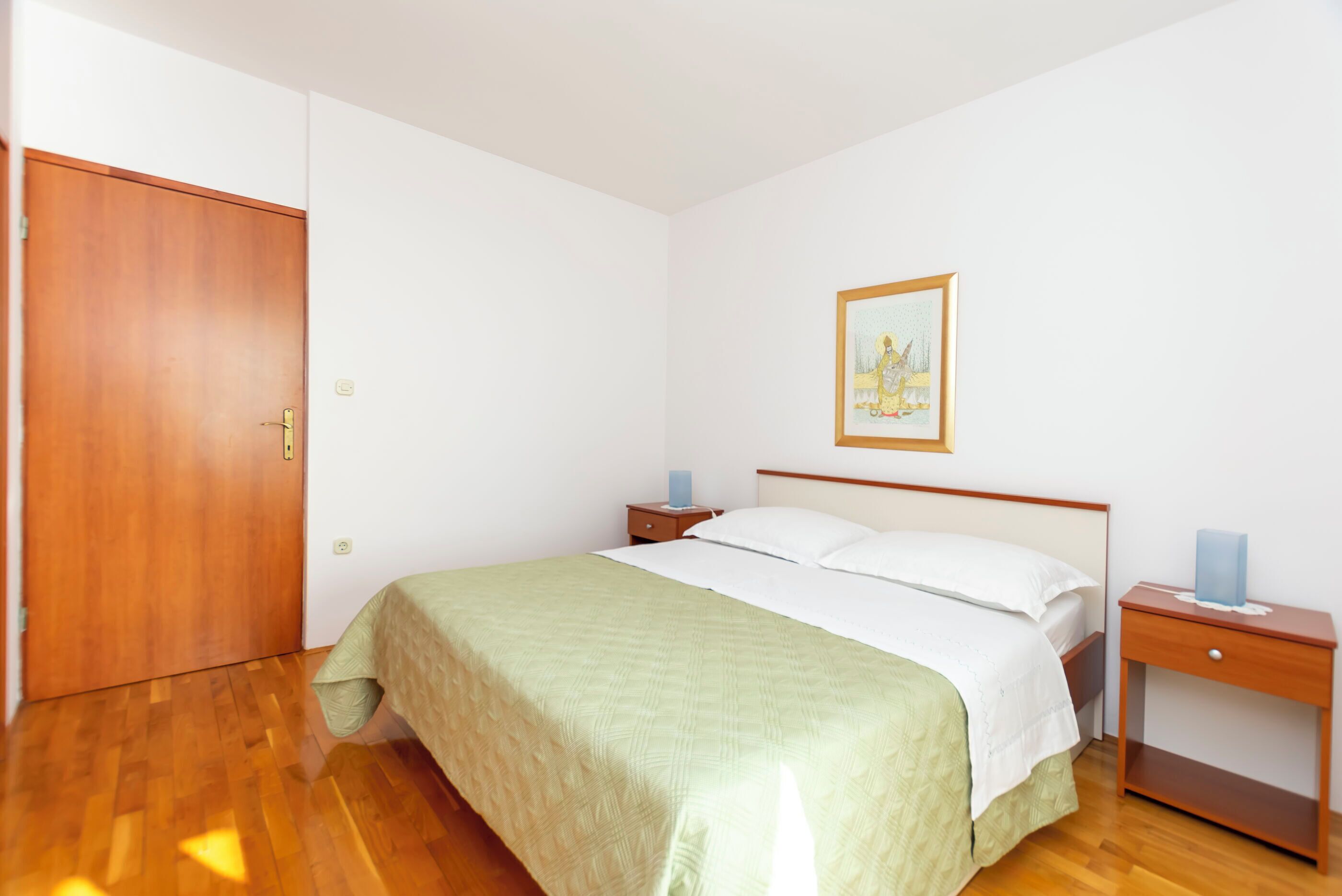 A bedroom with a wooden door, a bed with a green bedspread, two bedside tables with lamps, and a framed picture on the wall.