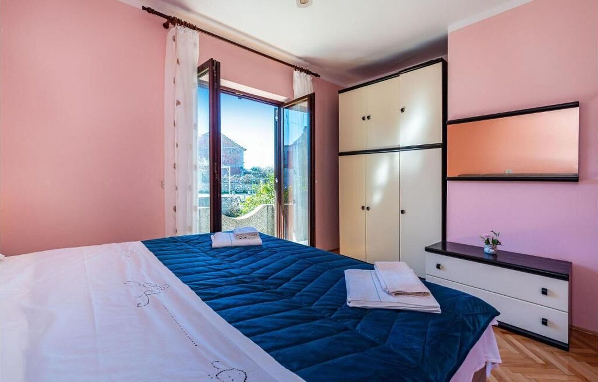 Ein Schlafzimmer mit Bett, Kleiderschrank, Kommode und einem großen Fenster mit Blick ins Grüne.