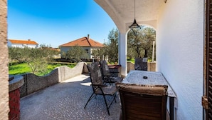 A covered outdoor patio with a table and chairs, a hanging light fixture, and a view of a house with a red roof in the background. - Apartment Serda (Preko)
