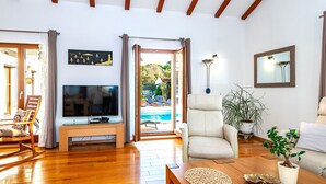 A spacious living room with a wooden ceiling, a television, a comfortable chair, a coffee table, and a view of an outdoor swimming pool. - Villa Pamaroka (Sibenik)