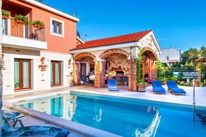 A swimming pool with lounge chairs, a covered patio area, and a building with a red-tiled roof. - Villa Pamaroka (Sibenik)