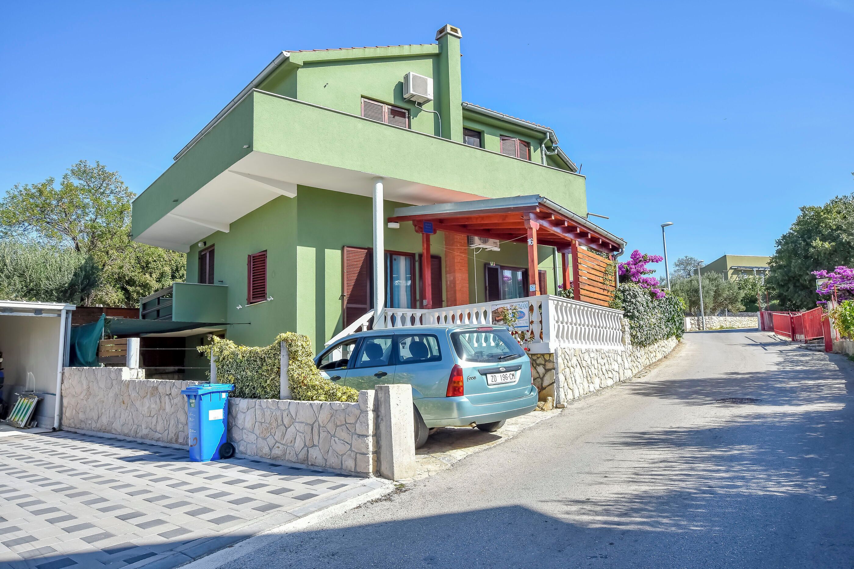 A two-story house with a balcony, a parked car, and a stone wall.