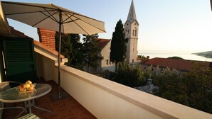 Ein Balkon mit Tisch und Stühlen, einem großen Sonnenschirm und Blick auf eine Kirche sowie die Küste.