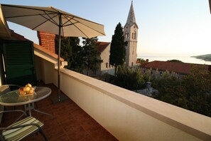 Ein Balkon mit Tisch und Stühlen, einem großen Sonnenschirm und Blick auf eine Kirche sowie die Küste.