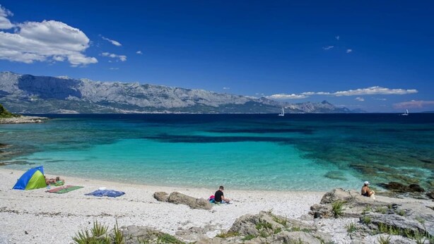 Ein Strand mit klarem türkisfarbenem Wasser, felsigem Ufer und Bergen im Hintergrund.