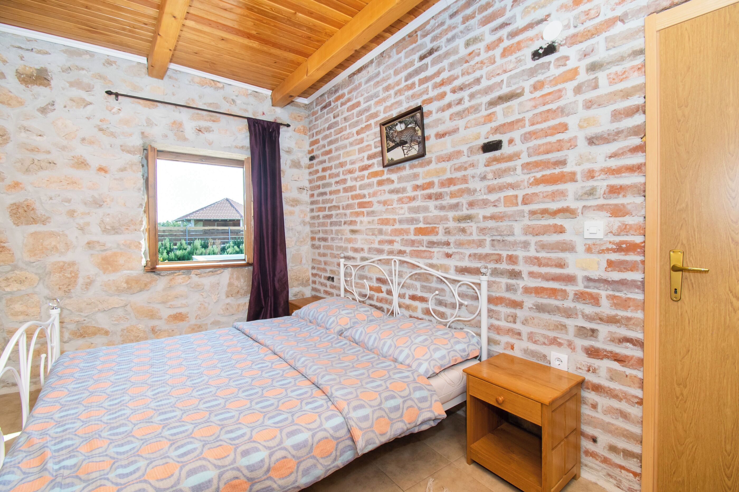 Une chambre avec un mur en briques, un lit avec une literie à motifs, une fenêtre donnant sur un paysage agréable et une porte en bois.
