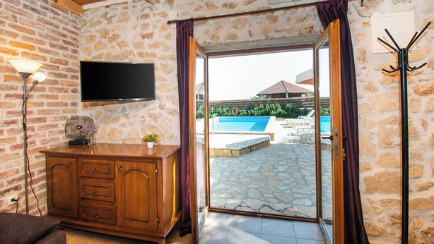 A room with a stone wall, wooden ceiling, a TV mounted on the wall, a wooden cabinet, and a sliding glass door leading to an outdoor swimming pool area. - House Stone Curlew 2 (Benkovac)