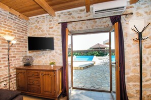 A room with a stone wall, wooden ceiling, a TV mounted on the wall, a wooden cabinet, and a sliding glass door leading to an outdoor swimming pool area. - House Stone Curlew 2 (Benkovac)