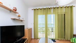 A modern living room with a flat-screen TV, wooden shelves, a glass table, and a view through green curtains to an outdoor area. - Apartments Luci (Rab)