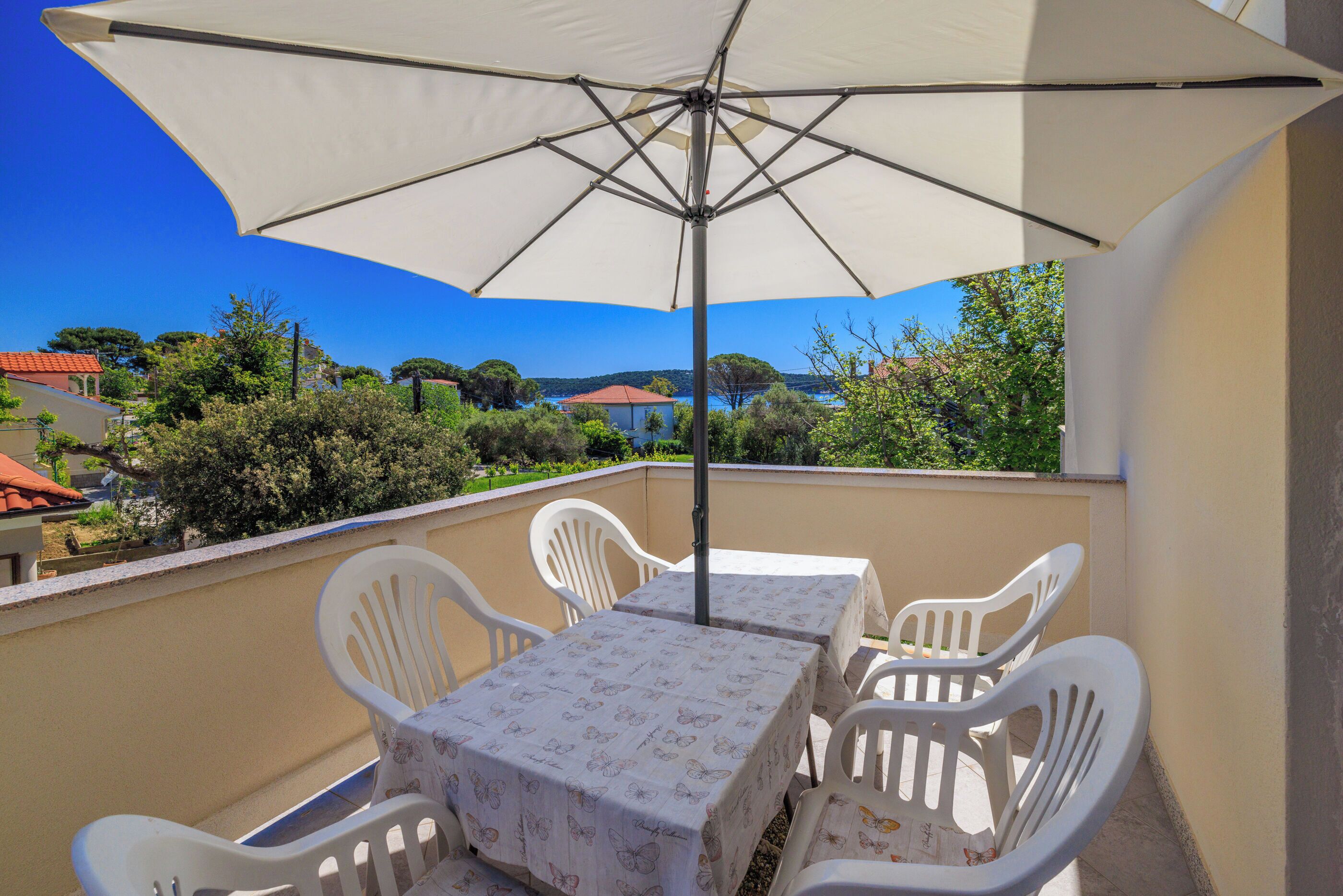 Un balcon avec une table et des chaises, protégé par un grand parasol, d’où l’on peut admirer un paysage pittoresque composé de maisons et d’arbres.