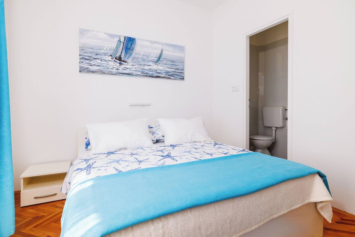 A bedroom with a bed, a painting of sailboats, a nightstand, and a bathroom door.