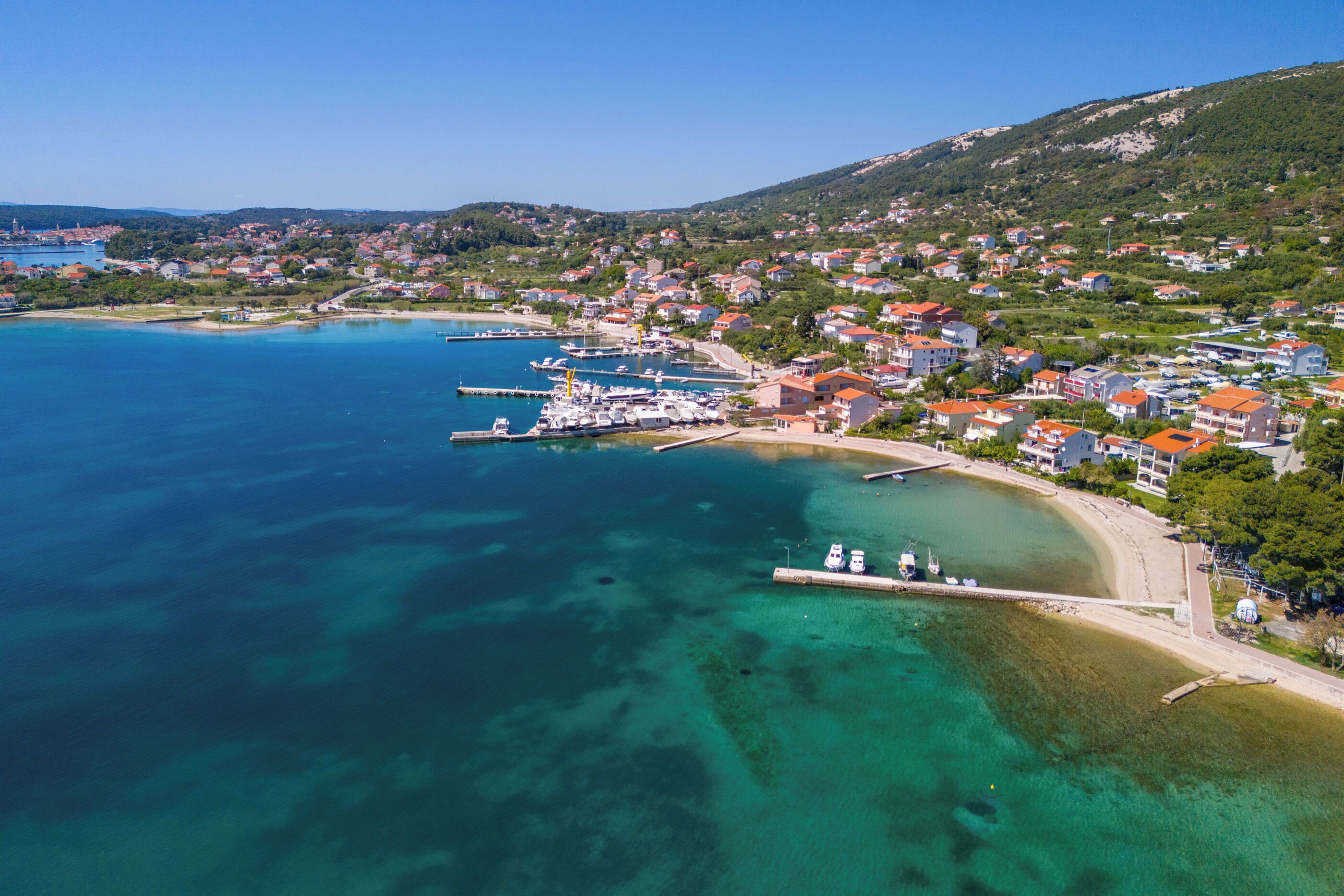 An aerial view of a coastal town with clear blue waters, a marina, and residential buildings.