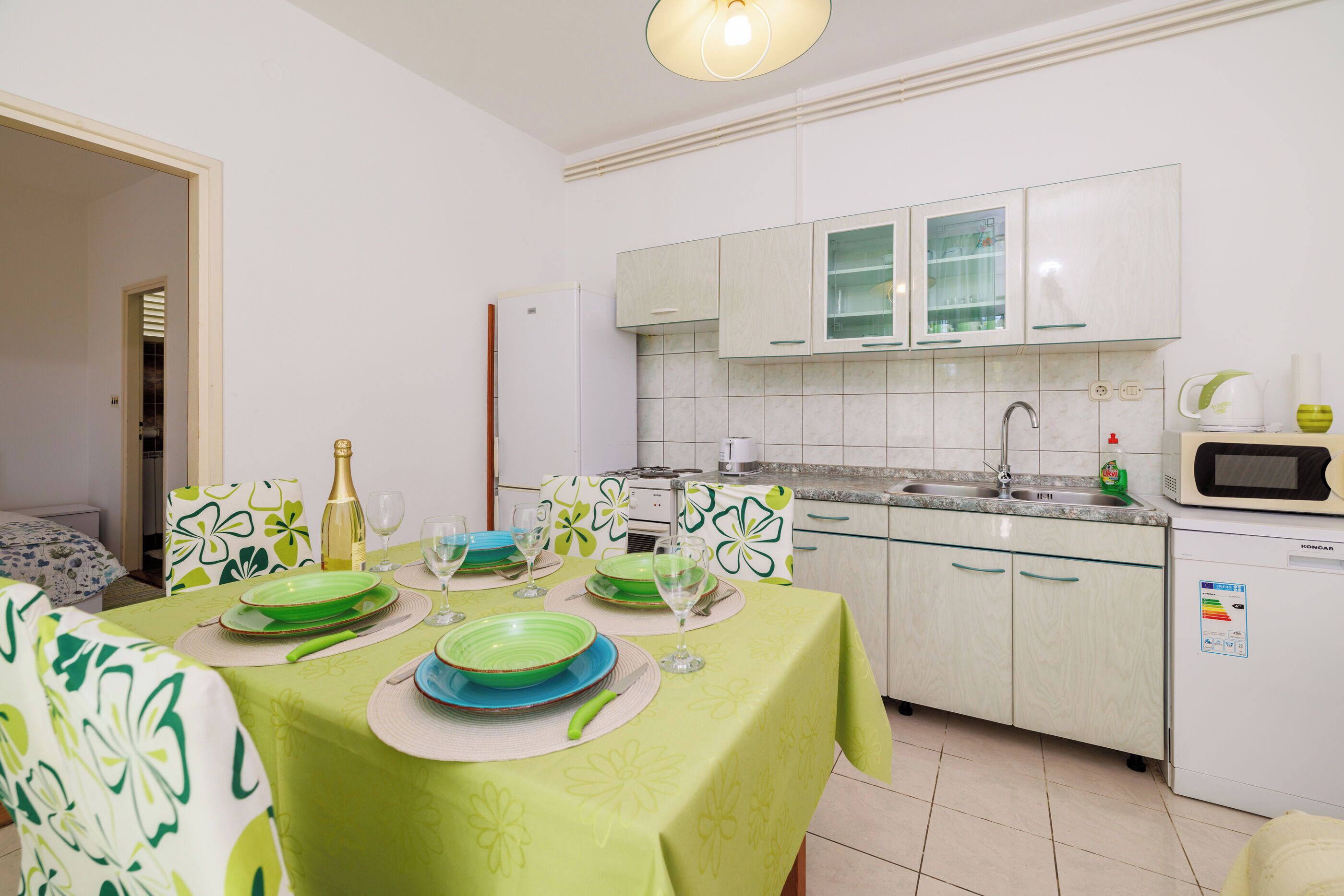 Een moderne keuken met witte kastjes, een magnetron en een vaatwasser. Er staat een eettafel gedekt met groene borden en glazen.