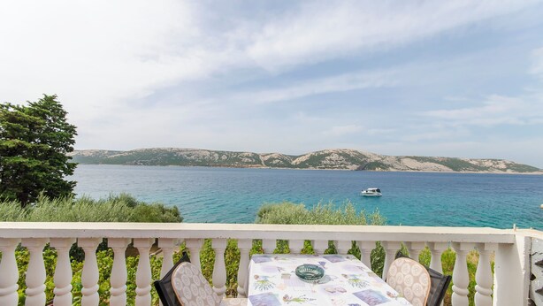 A balcony with a table set for two, overlooking a sea with a boat in the distance and a mountainous coastline. - Apartments Seafront Jadranka (Novalja)