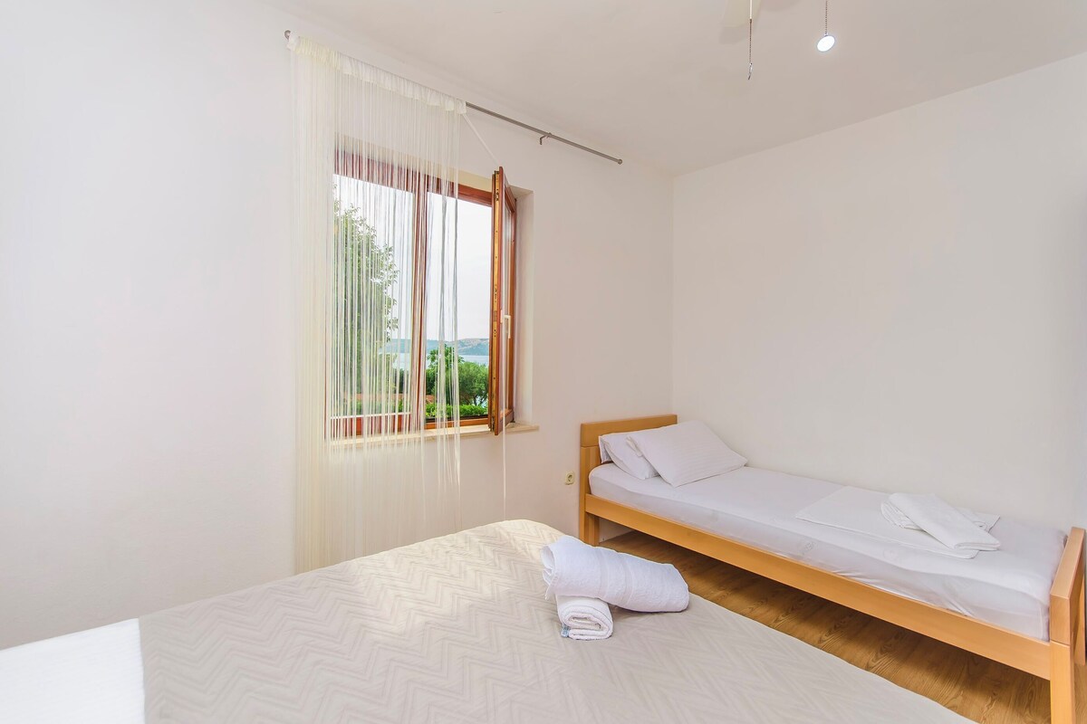 A clean, minimalist bedroom with a bed, a window with sheer curtains, and a view of greenery outside.