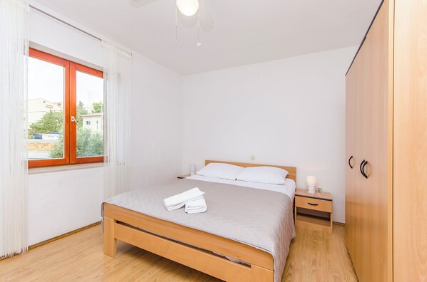 A bedroom with a wooden bed, a bedside table with a lamp, a wardrobe, and a window with curtains. - Apartments Seafront Jadranka (Novalja)