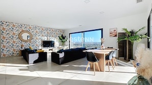 Uma sala de estar moderna com uma janela ampla, uma televisão de tela plana, um sofá, uma mesa de jantar com cadeiras e uma planta decorativa.