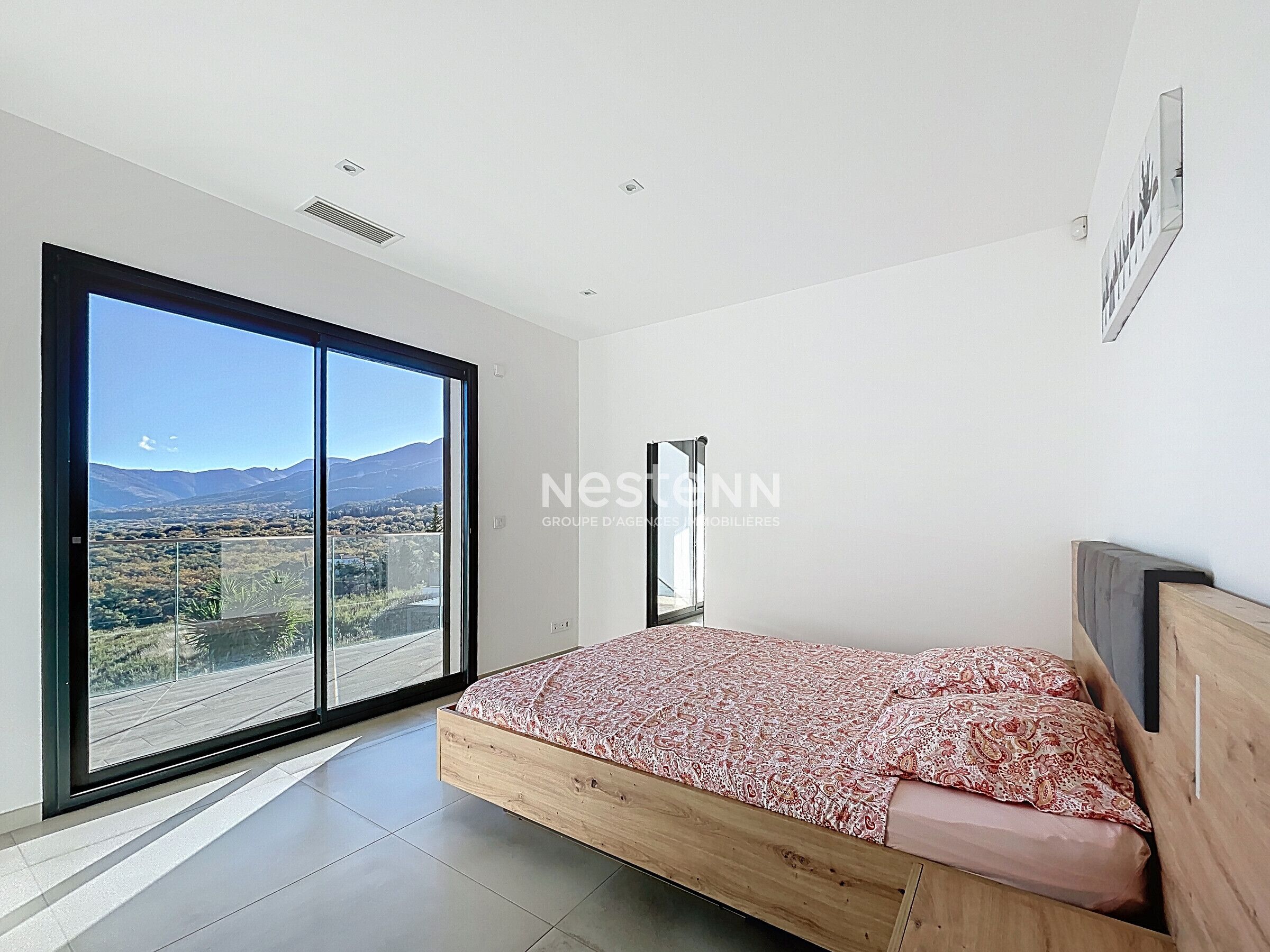 A bedroom with a large sliding glass door leading to a balcony, a bed with a patterned bedspread, and a mounted mirror on the wall.