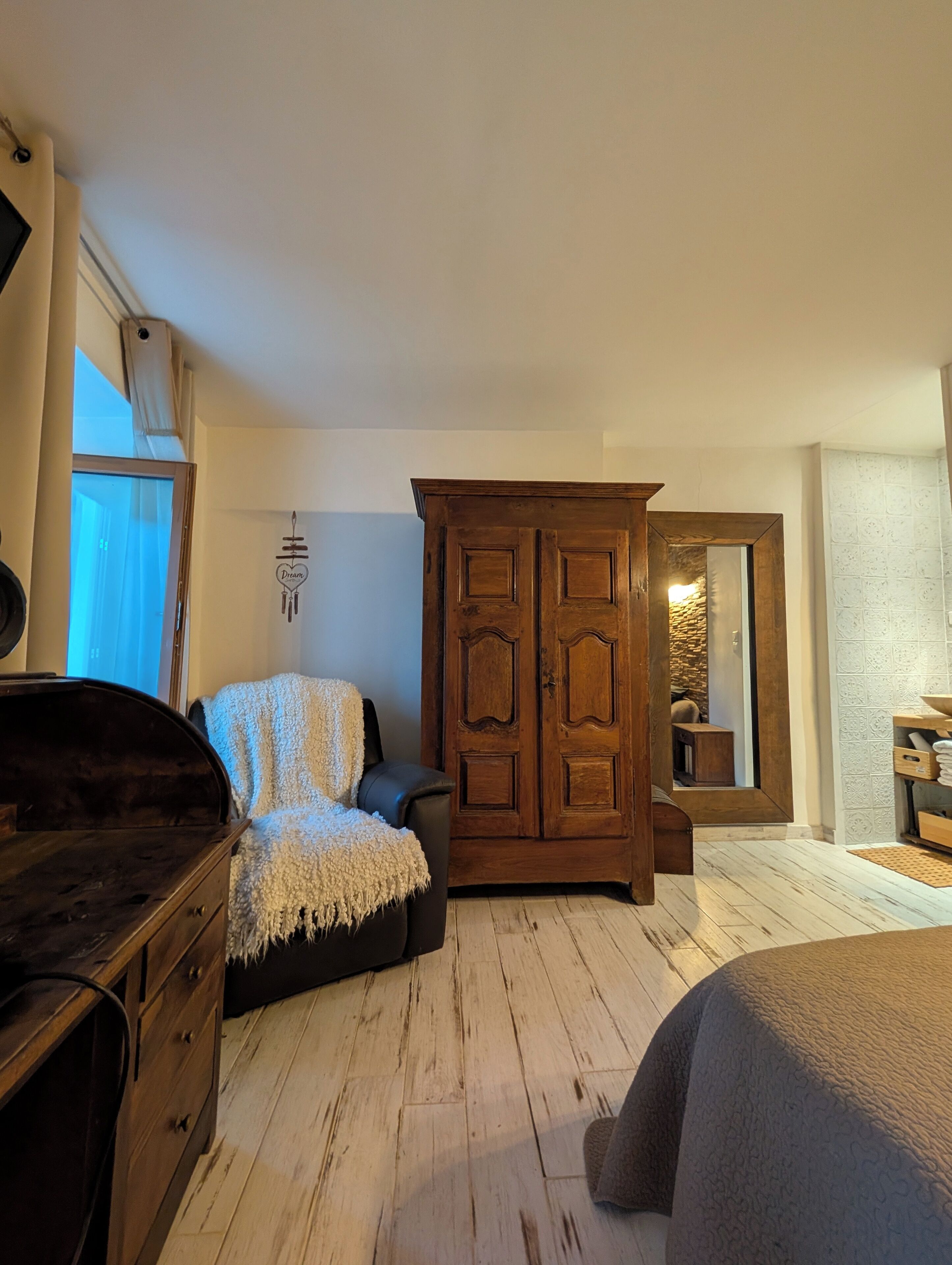 Ein Schlafzimmer mit einem Holfischschrank, einem Bett mit grauer Decke, einer Holzkommode und einem Fenster mit Vorhängen.