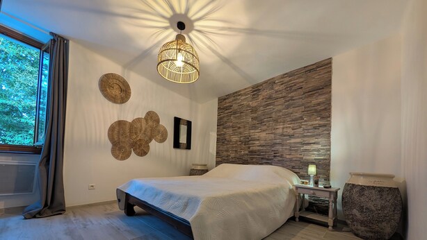 A bedroom with a bed, a bedside table, a lamp, a decorative wall, and a window with curtains. - Maison Cévenole - Village center - Flexible arrival (Saint-Jean-du-Gard)