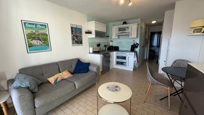 Un espace de vie compact comprenant un coin cuisine, un canapé gris avec des coussins, une petite table ronde et une chaise.