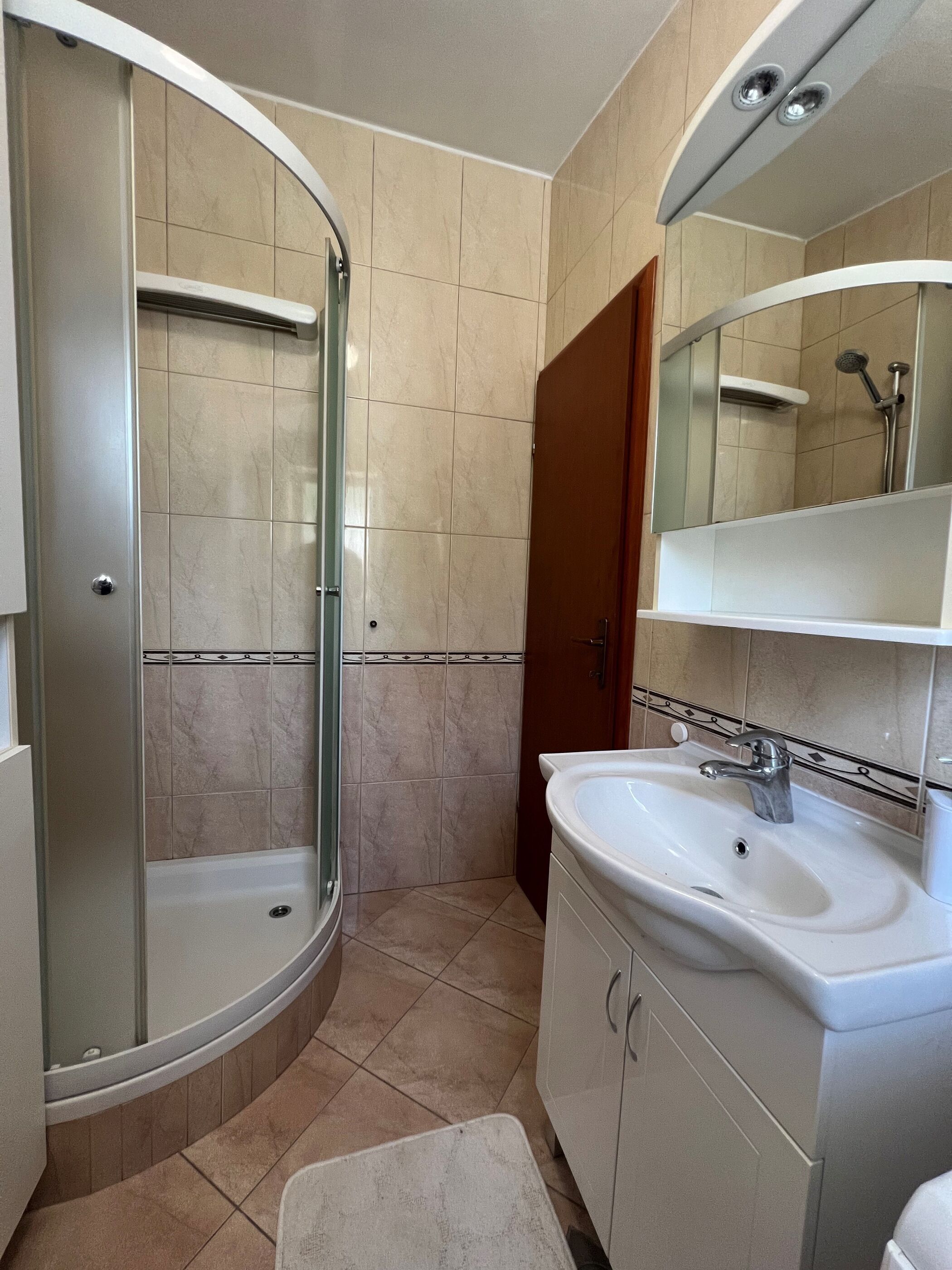 Un baño con ducha separada por mampara de vidrio, lavamanos con espejo y paredes con azulejos color crema.