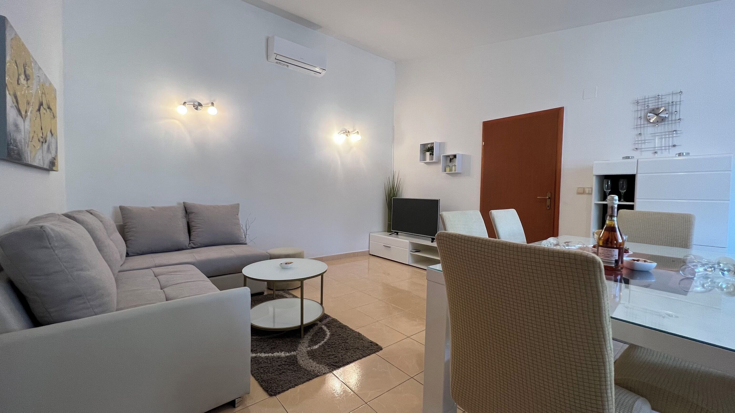 Ein modernes Wohnzimmer mit einer Couch, einem Fernseher, einem Esstisch mit Stühlen und einer Küchenzeile mit Kühlschrank.
