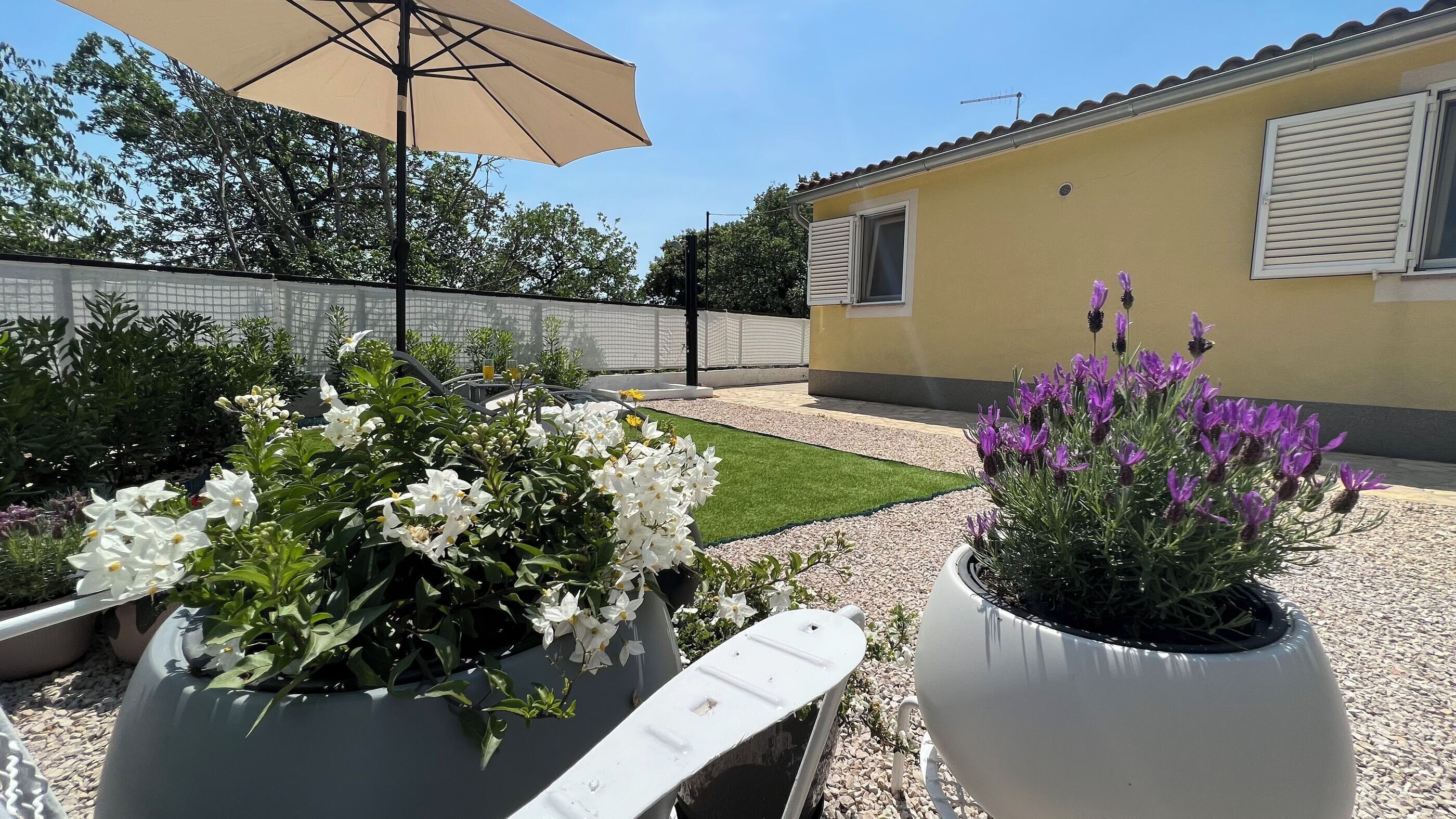 A garden with white and purple flowers, a white planter, and a beige umbrella.