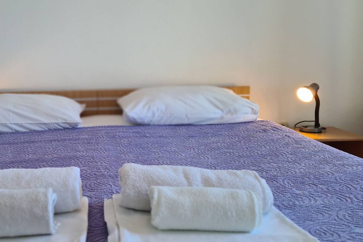 A neatly made bed with a purple patterned bedspread, two white pillows, and two rolled towels on the bed.