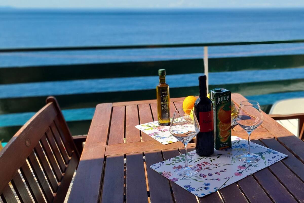 A wooden table with two wine bottles and glasses, set on a floral placemat, overlooking the sea.
