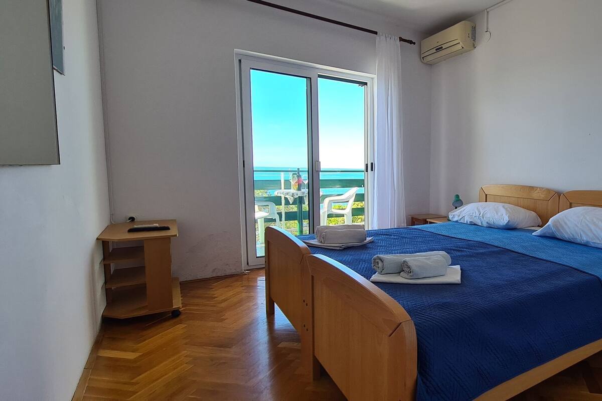 A bedroom with a bed, a nightstand, a desk, and a view of the sea through a sliding door.