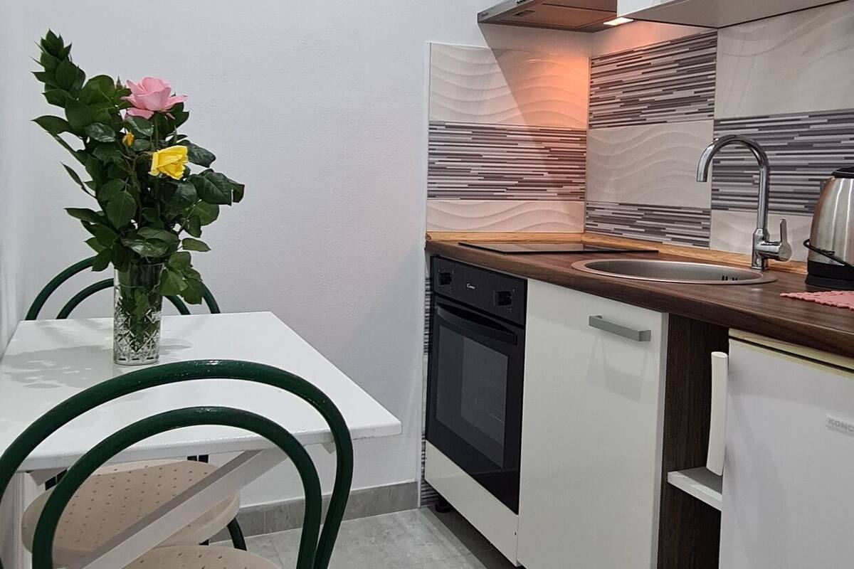 A compact kitchen with white cabinets, a built-in oven, and a sink. There is a dining table with chairs and a vase of flowers.