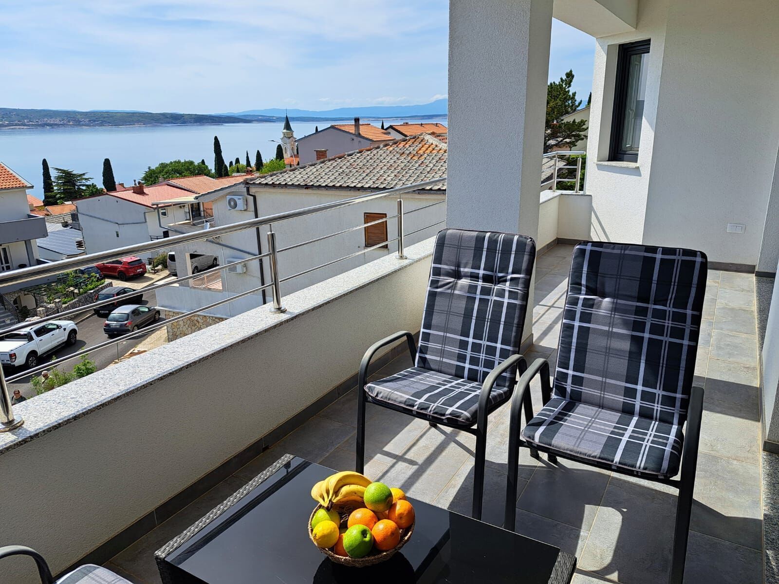 A balcony with two chairs and a table, a bowl of fruit, and a view of a lake and buildings.