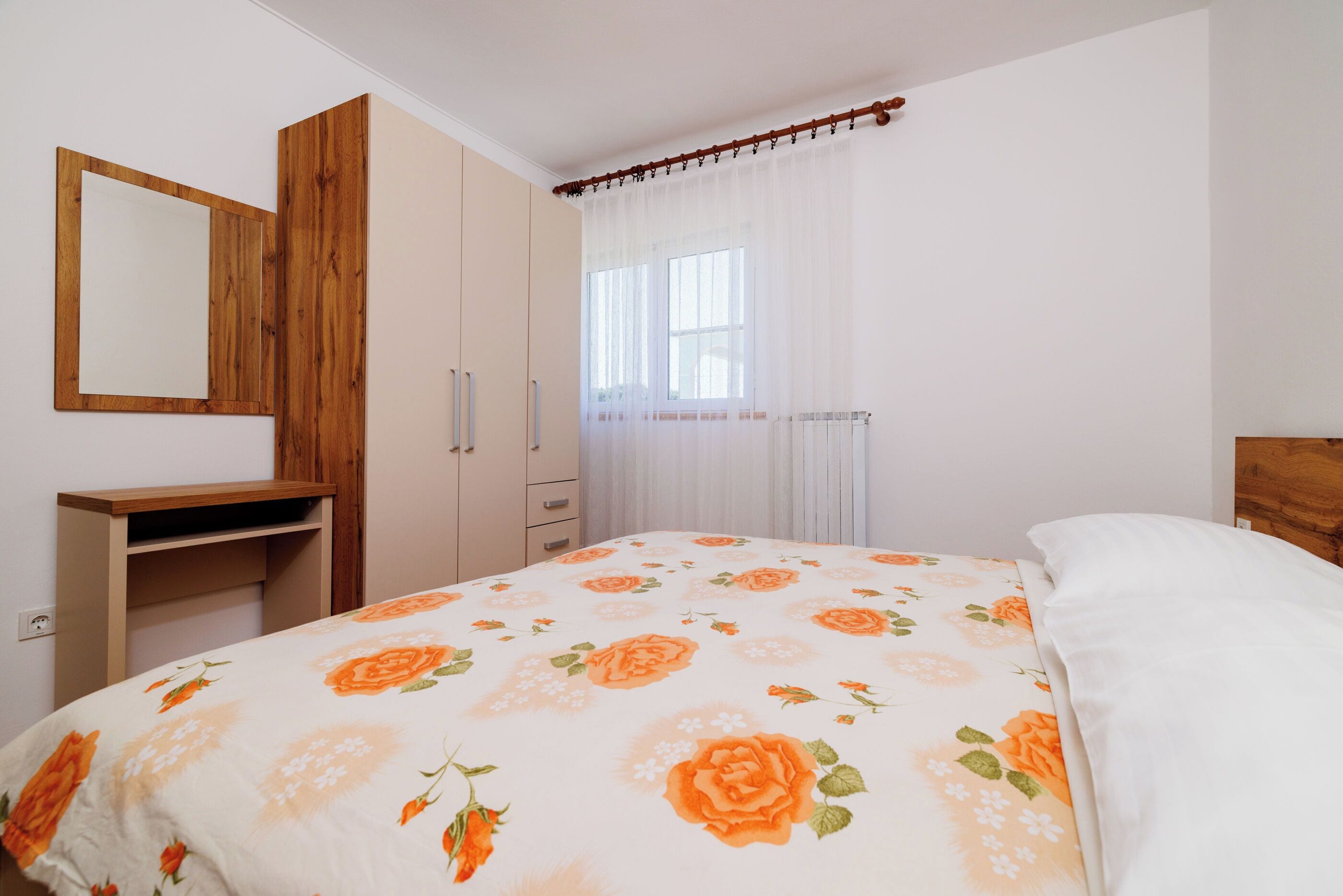 A bedroom with a bed, a wooden wardrobe, a mirror, and a window with sheer curtains.