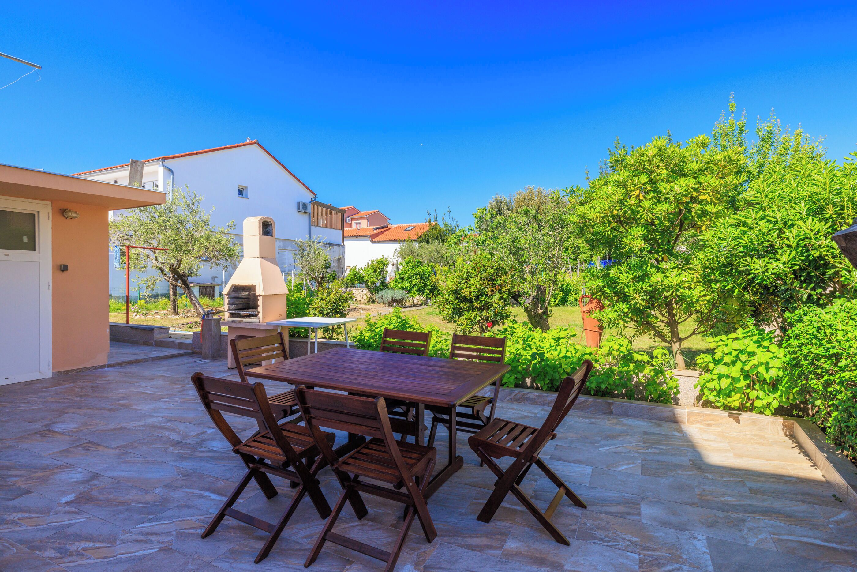 Un patio con tavolo e sedie in legno, circondato da pavimento in pietra, con vista su un edificio bianco e vegetazione sullo sfondo.