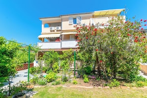 Un immeuble résidentiel à plusieurs étages, doté de balcons, d’un jardin avec différentes plantes et d’un arbre aux fleurs rouges, le tout sous un ciel bleu clair.