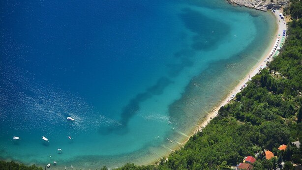 Vue aérienne d’une zone côtière avec des eaux d’un bleu limpide, une plage de sable et une petite péninsule rocheuse à la végétation clairsemée.