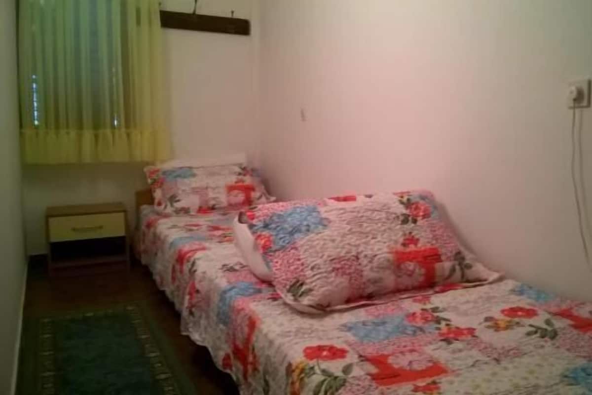 Une petite chambre agréable et chaleureuse, avec deux lits simples, chacun agrémenté d’une literie à motifs floraux. On y trouve une petite table de chevet en bois avec une lampe, ainsi qu’une fenêtre avec des rideaux.