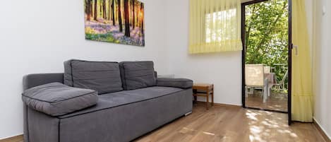 A modern living room with a grey sofa, a painting of a forest, a wooden floor, and a sliding glass door leading to an outdoor area.