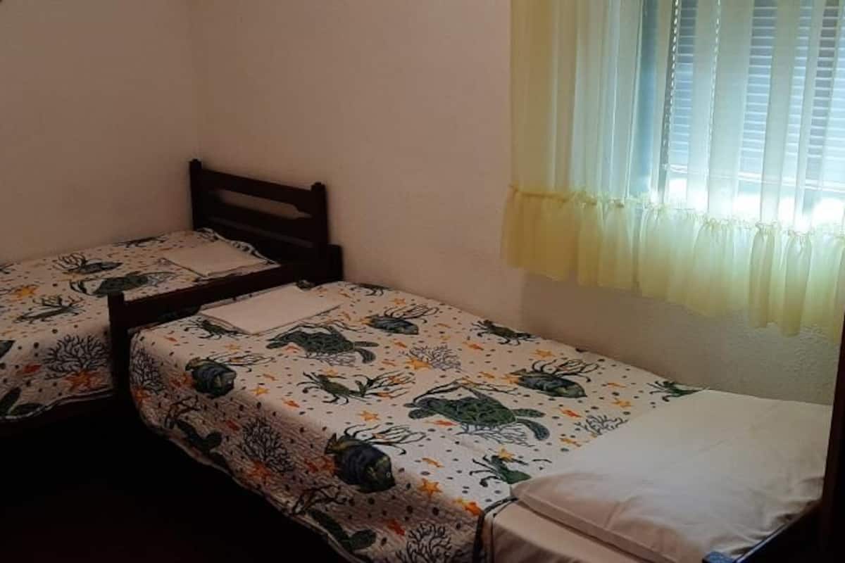 Une petite chambre avec deux lits simples, chacun recouvert d’une couvre-lit à motifs floraux. Il y a une fenêtre avec des rideaux légers et une tête de lit en bois.